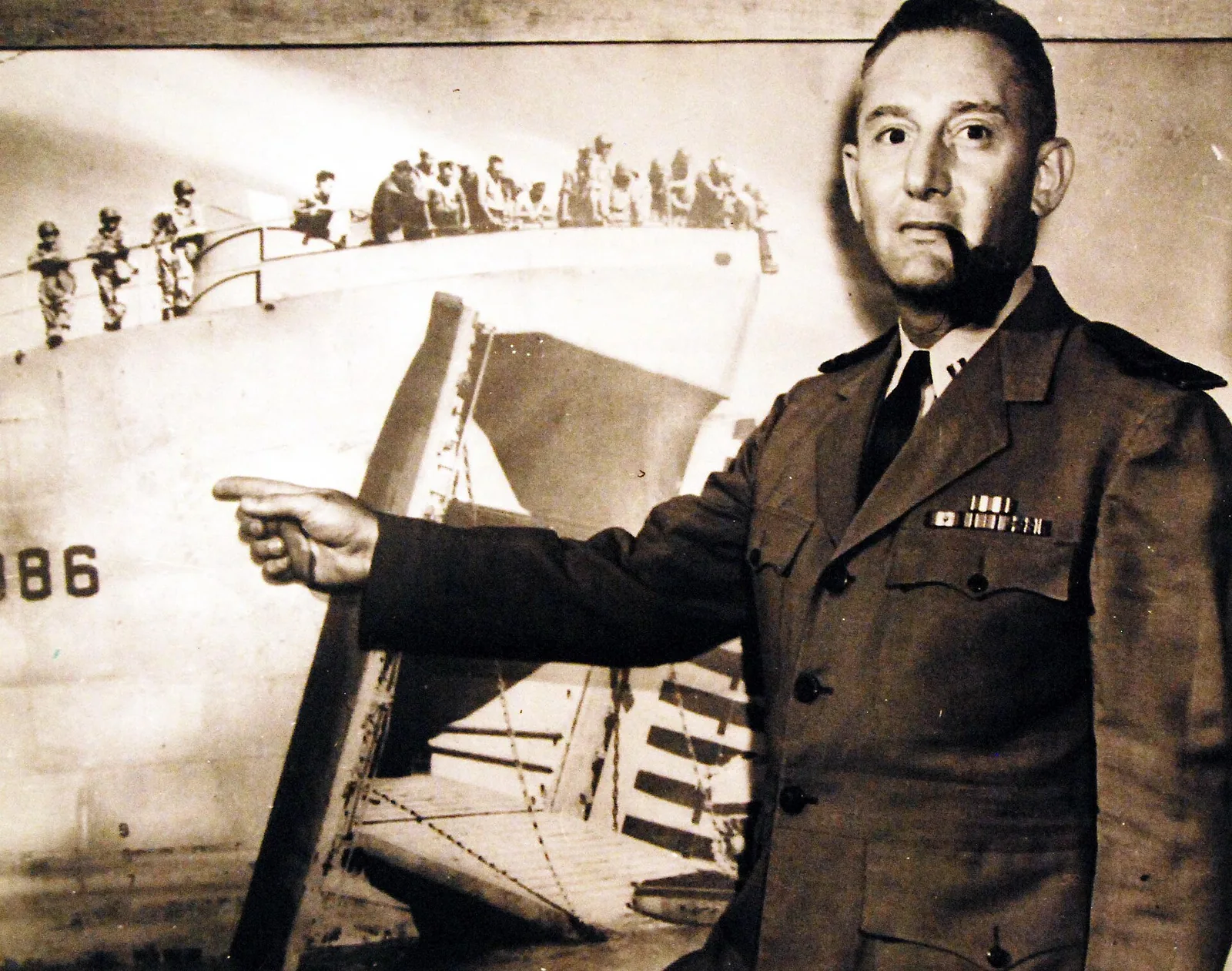 80-G-46566: Lieutenant Harold R. Fleck, USNR, poses against a photograph of his ship USS LST 386, as he recalls how the giant landing craft underweight the experiments which made it the first “junior aircraft carrier.” T…